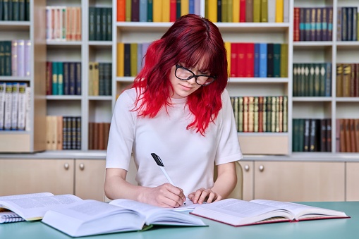Teenage female student studying in library class, sitting at desk using books, writing in notebook. Fashion student with red dyed hair wearing glasses. College, study, education knowledge, adolescence
