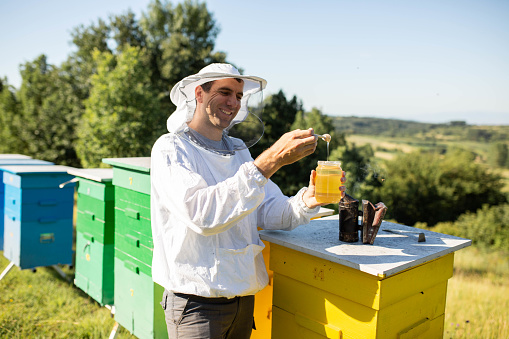 Portrait of a handsome beekeeper in protective uniform standing with honey in the jar, tasting fresh product on the apiary outdoors
