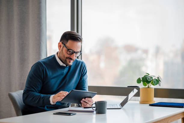 Young businessman using digital tablet while working in business office. Young businessman using digital tablet while working on laptop in business office. Male professional with wireless computer at desk. He is sitting by window in office. business person stock pictures, royalty-free photos & images