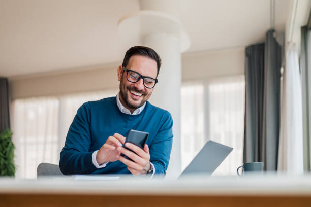 Small business entrepreneur looking at his mobile phone and smiling, reading or texting good news. Smiling businessman using phone in home or office. Small business entrepreneur looking at his mobile phone and smiling reading or texting good news. mid adult men stock pictures, royalty-free photos & images