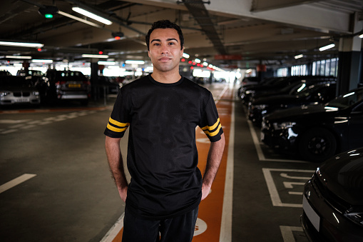 Portrait of young man standing in a car park and looking at camera. His outfit is black.