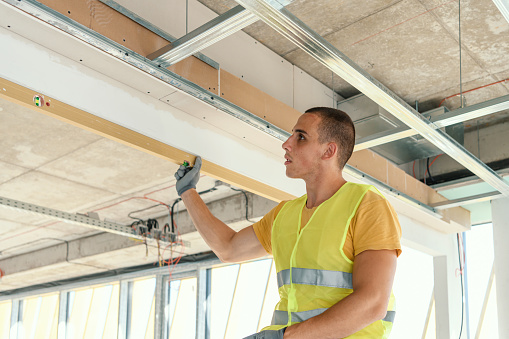 A young Caucasian male construction worker wearing gloves is using a level on a part of the ceiling.