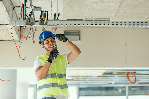 A young Caucasian male construction worker is cutting cables using pliers with a smile on his face.