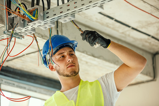 A young Caucasian male construction worker is handling the electrical wires with a serious look on his face.