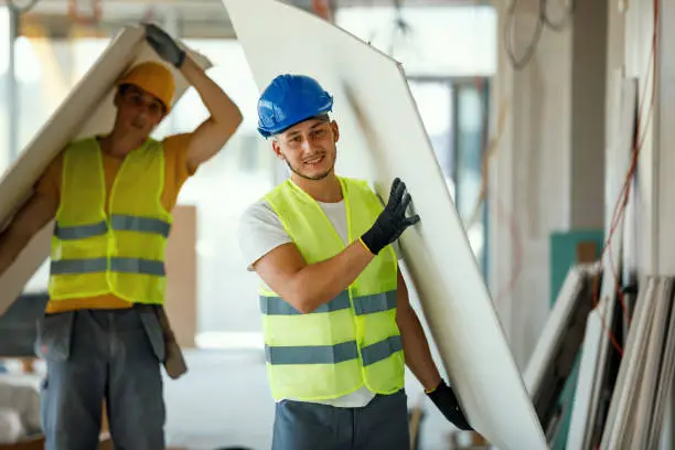 Two young Caucasian male construction workers are cheerfully carrying together pieces of drywall.