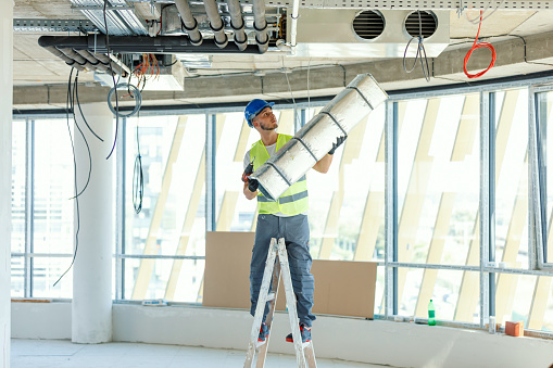 A young Caucasian construction worker is standing on a ladder and installing an air conditioner.