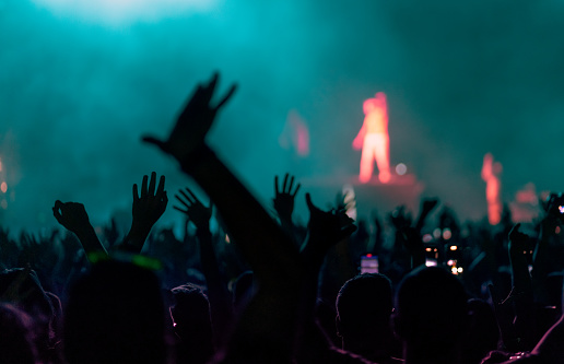 Rear view of a large group of people in front of a music festival stage. Crowd is excited and dancing, raising hands, clapping, punching the air, filming with mobile phones, etc... under teal stage lights.