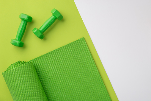 Fitness accessories concept. Top view photo of green sports mat and dumbbells on bicolor green and white background with copyspace