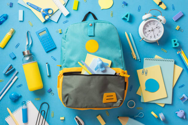 istock Back to school concept. Top view photo of open blue rucksack with plane shaped sharpener sticky note paper pens scattered school accessories drink bottle and alarm clock on isolated blue background 1409757196