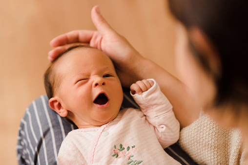 Cute funny infant baby girl making faces lying on mother hands in room at home and try to fall asleep. Motherhood. Woman holding newborn