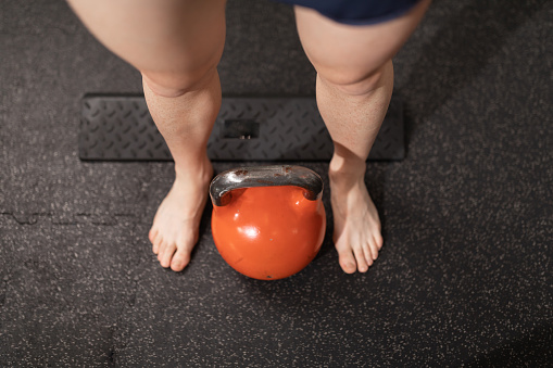 A kettlebell on the ground, between the bare feet of a young man resting after doing sport at the local training and fitness center