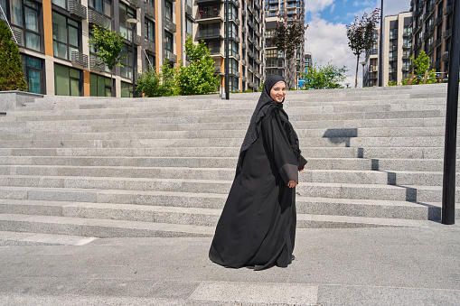 Muslim woman in a veil stands on the stairs near high-rise buildings
