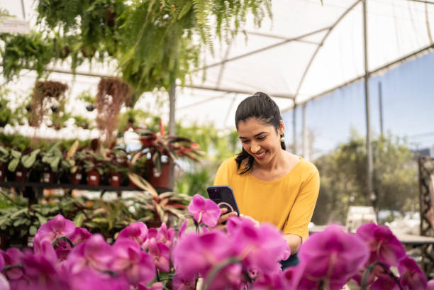 jovem fotografando ou filmando flores no celular em um centro de jardim - garden center flower women plant - fotografias e filmes do acervo