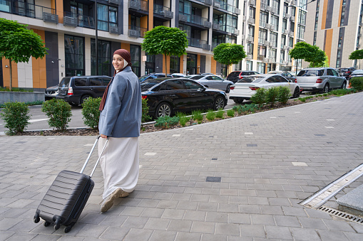 Woman in a hijab stands on the street in a half turn and holds a suitcase on wheels