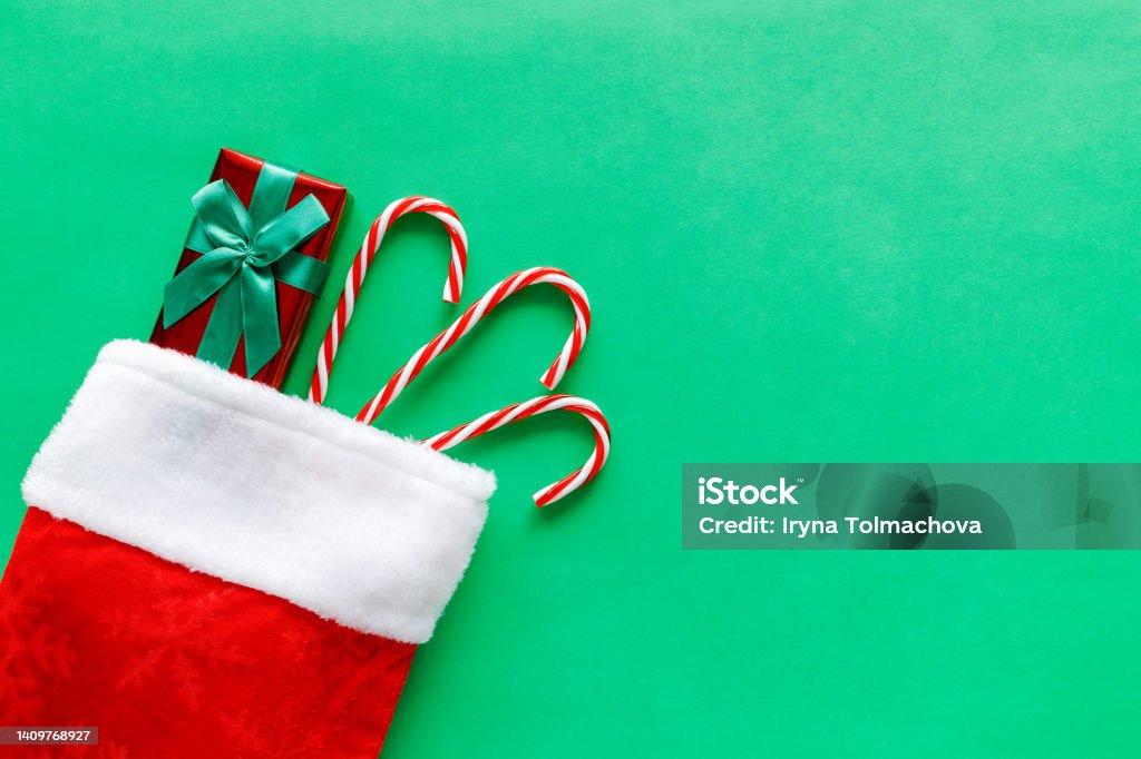 Christmas stocking with gift box and candy canes on green background. Christmas stocking with small gift box and candy canes on green background with copy space for text. Flat lay composition of holiday decorations. Christmas Stocking Stock Photo