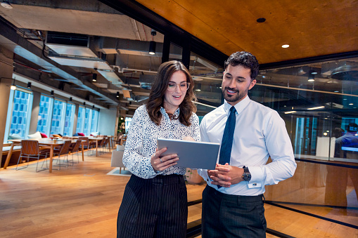 Business Man and woman standing working together on a digital tablet. They are both well dressed standing in a modern office. They are happy and smiling