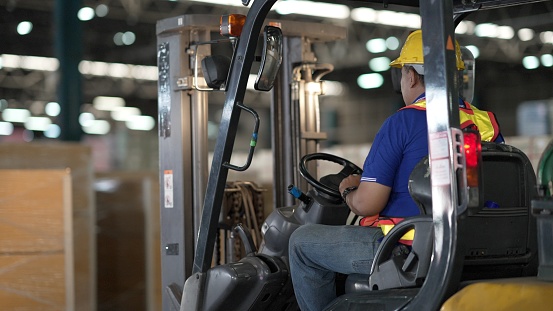 Rear view. Electric forklift truck operator drive and lifts pallet with carton boxes on a shelf. Employee working, scanning products, using trucks in distribution center or retail warehouse full of shelves with goods. Concept of transportation, supply chain.