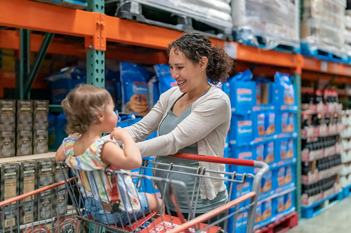 A multiracial woman of Pacific Islander descent who is pregnant sweetly interacts with her toddler daughter who is sitting in a shopping cart as they grocery shop together.