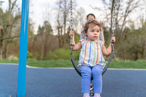 A cute multiracial toddler girl sits on a swing at the park while enjoying time outside with her mom,