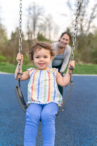 A gorgeous Eurasian toddler girl smiles directly at the camera as her pregnant mom pushes her on a swing at the park.