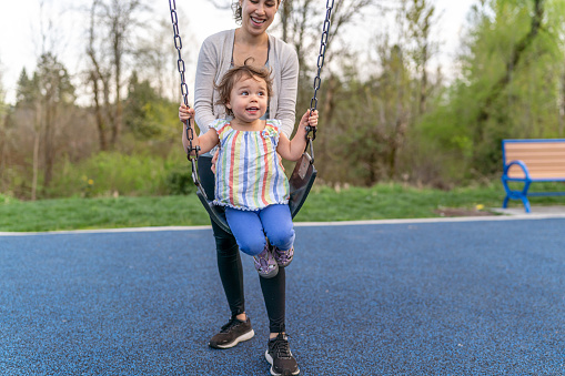 An adorable and happy Eurasian toddler girl looks excited as her beautiful Pacific Islander mom pushes her on a swing at the park.