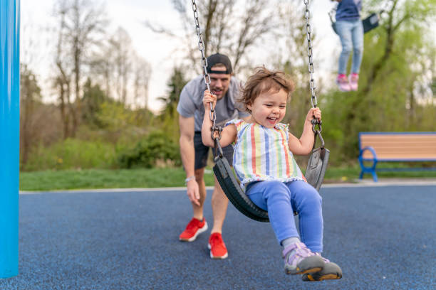 An adorable and happy Eurasian two year old girl laughs as her dad pushes her on a swing at the park.