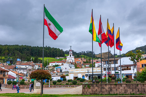 Zipaquira, Colombia - June 30, 2016: Looking across Independence Square in the Andean city of Zipaquira, in the South American country of Colombia towards the chapel on the hill on an overcast day. The town is located on the Eastern Range of the Andes Mountains, at an altitude of 8,690 feet above mean sea level. The present day city was established in the year 1600 AD. Spanish colonial architecture and it's influence can be seen in the image. Zipaquira's flag can be seen to the left, separated from the other flags, which include the Colombian National flags. Photo shot in the afternoon sunlight on an overcast day; horizontal format.