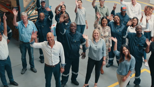 Portrait of a diverse group of employees waving and smiling in an industrial warehouse from above. Engineers and office staff workers smile and do friendly greeting gestures in a mechanics workshop