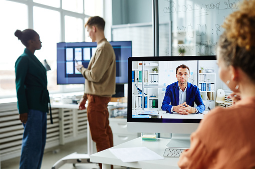 Woman sitting at her workplace and talking to her colleague on computer monitor during video call at office