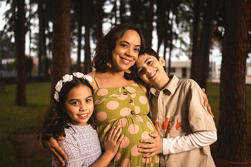 pregnant latin mom hugging her son and daughter in pine forest looking at the camera