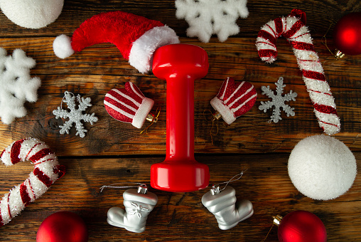 Red dumbbell stylized as Santa Claus. Hat on top. Hands, legs arranged with glove, boot Christmas decorations. Snowflakes, candy canes ornaments around. Healthy fitness lifestyle holiday season concept. Fit gym composition on wooden background.