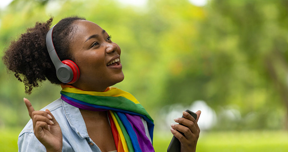 African American LGBTQ+ woman is listening to music from her smartphone while sitting relaxingly in the public park during summer