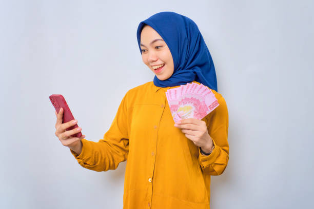 aufgeregte junge asiatische muslimische frau in orangefarbenem hemd mit handy- und rupiah-banknoten, die isoliert über weißen hintergrund auf online-sieg reagiert - indonesian currency stock-fotos und bilder