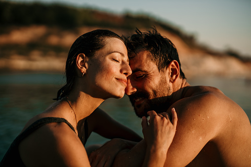 Affectionate couple sitting by the water in swimming suits, cuddling and enjoying the sunset after swimming in the sea. Couple on vacation concept