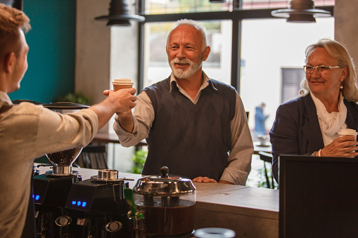 Candid shot of young male barista standing behind bar counter, handing a coffee, in a to do cup, to a senior man and his wife at the coffee shop.