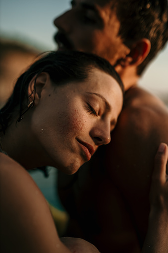 Sensational portrait of a lovable looking young adult European couple, enjoying wet, in their bathing suits, sitting next to the sea, hugging and cuddling. A beautiful brunet is leaning her head on a handsome's bearded partner shoulder. Both hugging, enjoying on the sun, feeling romantic
