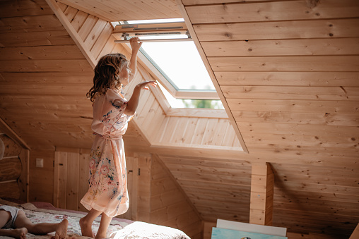 A little girl in a delicate pink dress is standing on the bed looking out the window. A child in a room under the roof of the house watches the birds through the window.