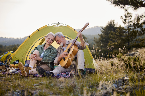 Cheerful mature man enjoying while playing a guitar to his wife during camping day in nature.