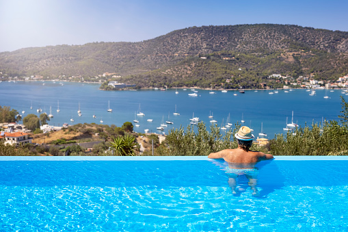 A tourist man in a swimming pool enjoys the view of the sailing boats in a bay at Poros island, Saronic Gulf, Greece, during summer time