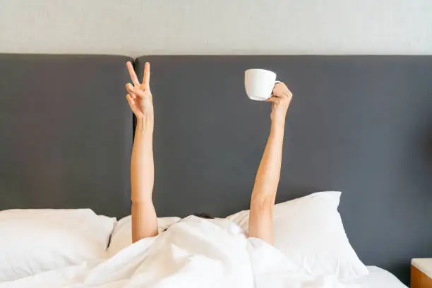 Photo of Young beautiful woman waking up in the morning in the bed, hiding under the blanket, stretching out her arms with a cup of coffee and showing V sign.