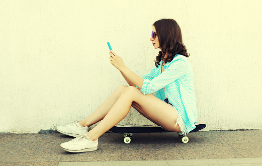Modern young woman with smartphone sitting on skateboard on city street on white background