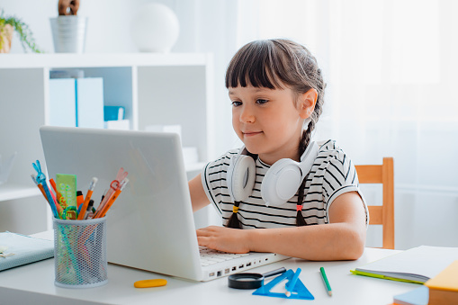 Child brunette schoolgirl studying homework during an online lesson at home in a bright white room, school time, online education concept, home school girl