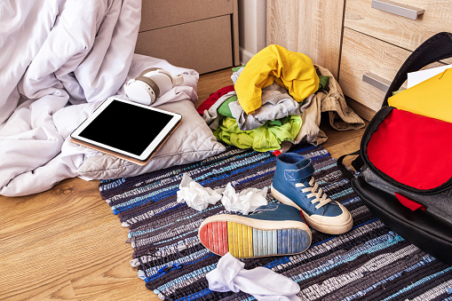 Colored clothes, sneakers, a backpack, a tablet, socks, an unmade bed are scattered on the floor in a mess in the room of a teenager who does not want to clean his room. Close-up, side view