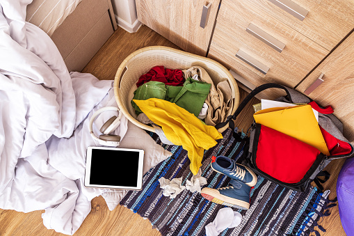 Top view, close-up. Colored clothes, sneakers, a backpack, a blanket, a briefcase are scattered on the floor in a big mess. The room of a teenager who does not want to keep order