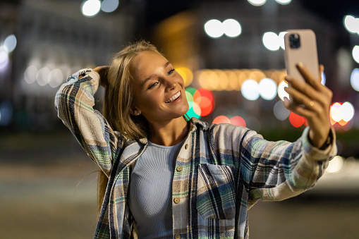Beautiful young woman taking selfie with a smartphone while walking on the city streets on the coast during the night