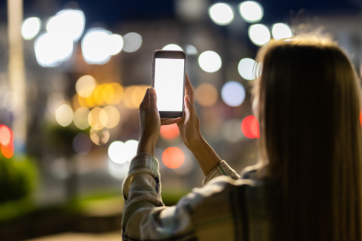 Young woman holding mobile phone with blank white display on background of light night street of the city