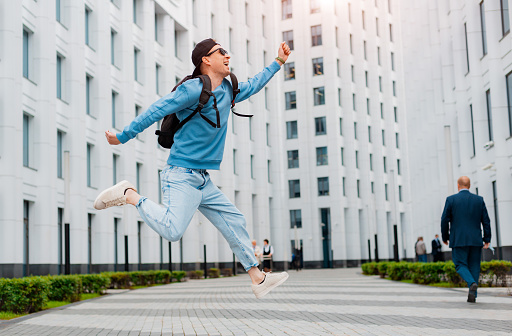 young stylish guy in blue clothes jumps on the background of a white modern building city street. .
