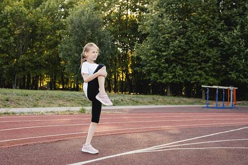 Child girl doing warm-up exercises on a sports track before running in a public park