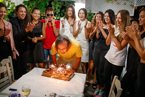 Man blowing out the candles on his birthday cake, his happy friends standing around him, singing and clapping hands with joy