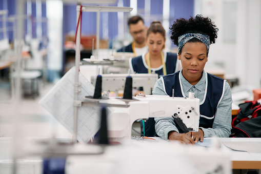 Young African American woman sewing while working as seamstress at clothing factory.